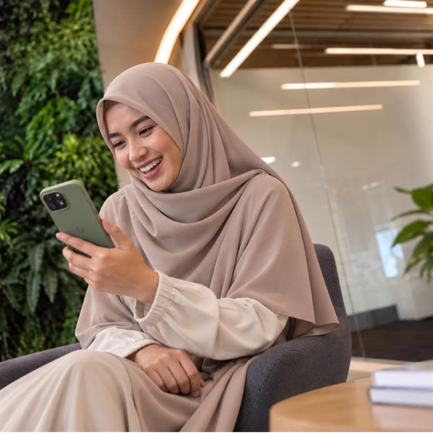 Perempuan berhijab tersenyum melihat smartphone di ruang santai